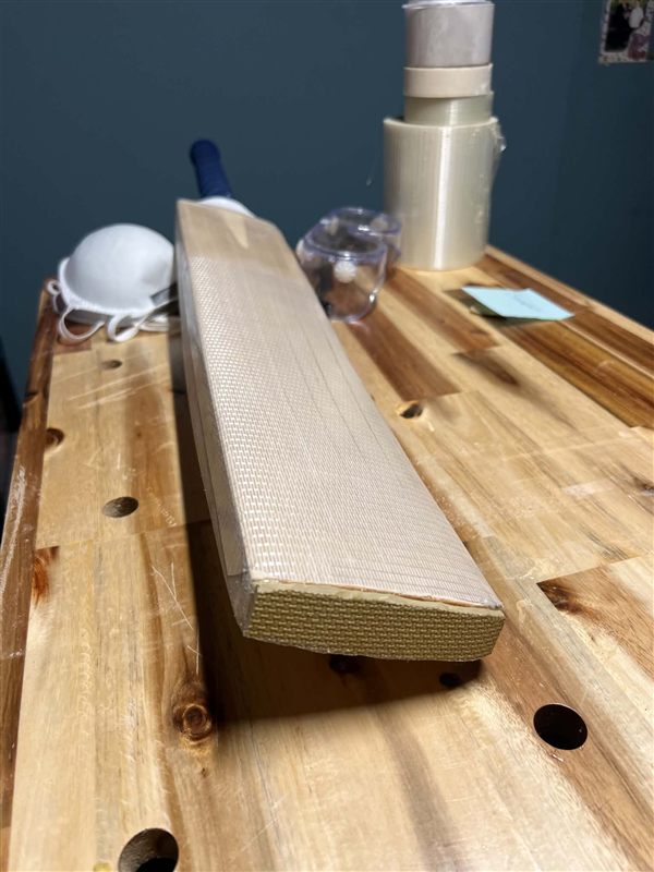 Bat lying on a workbench with fibreglass facing across the blade and a toe guard on the toe, rolls of facing tape behind